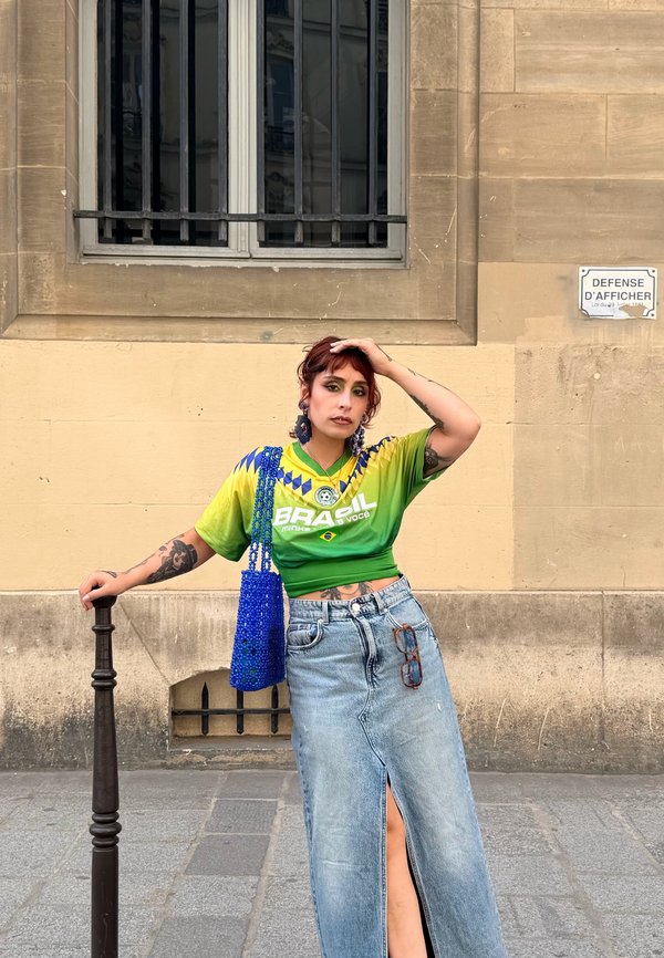 Jeune personne aux cheveux roux appuyée contre un poteau, portant un maillot de football vert du Brésil, une longue jupe en jean fendue, un sac bleu, debout devant un mur beige.