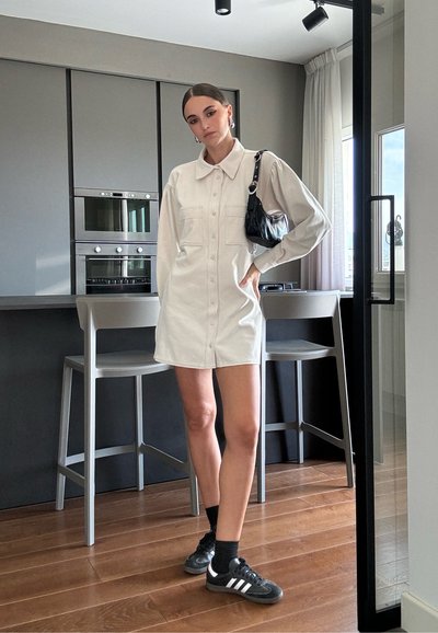 Vestido blanco de botón con dos bolsillos frontales, tejido suave, ajuste relajado. Combinado con un bolso negro y zapatillas negras de Adidas. Suelo de madera, cocina gris.