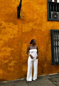 Weißes trägerloses Oberteil und weite Hosenbeine vor einer strukturierten orangefarbenen Wand. Auffällige Details sind große Creolen und geschichtete Armbänder.