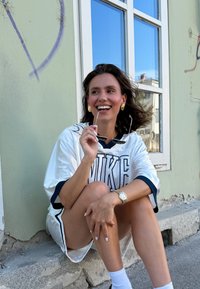 Fehér sportmez sötétkék részletekkel és „NIKE” felirattal, ezüst órával és oversize napszemüveggel párosítva. Kőfelszínen ül.