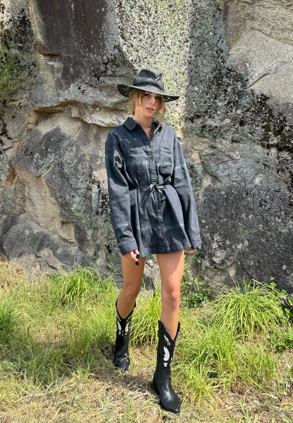 Vestito nero a camicia con chiusura a bottoni, legato in vita, abbinato a stivali da cowboy neri alti con accenti floreali bianchi. Sfondo in pietra naturale.