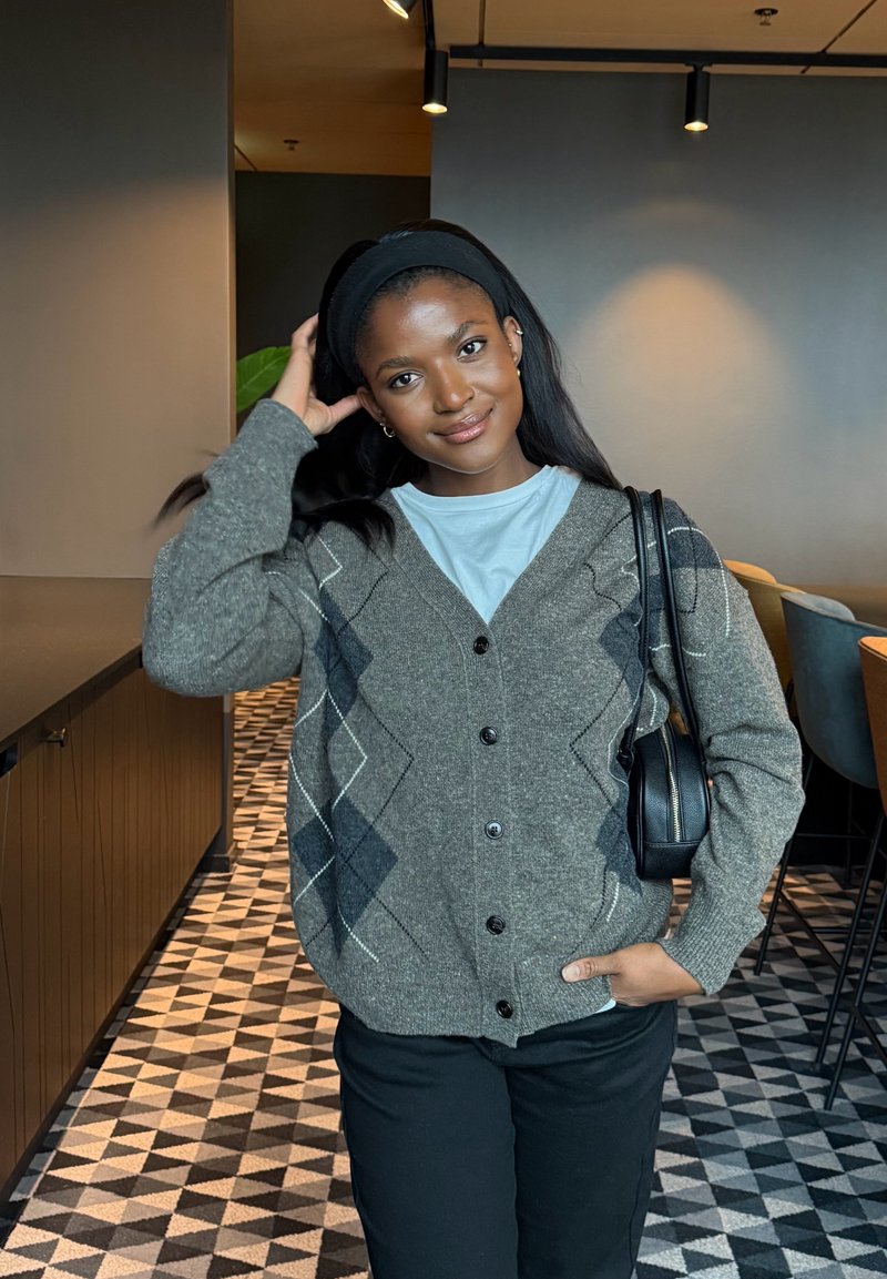 Giovane donna con capelli scuri che indossa un cardigan grigio a motivo rombi, camicia bianca e pantaloni neri, posa al chiuso con una mano sulla testa e l'altra in tasca.