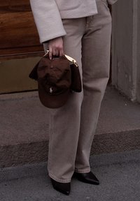 Personne portant un pantalon beige et des chaussures noires à bout pointu, tenant une casquette marron et un sac à main assorti, sur fond de rue et de bâtiments.