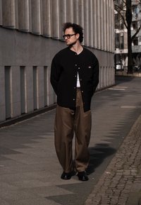 Casaco acolchoado preto com fecho de botão e bolsos laterais, usado sobre uma t-shirt branca. Calças largas bege e sapatos pretos. Fundo urbano.