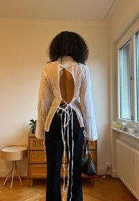 White long-sleeve shirt with a large back cut-out, tied with ribbon lacing. Paired with black trousers and a small black bag.
