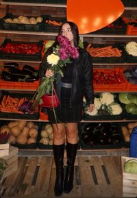Veste en cuir noire, robe à motifs, bottes noires montant jusqu'aux genoux ; tenant des fleurs roses et blanches devant un fond de légumes colorés.
