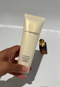 Nettoyant en crème mousse MAC Hyper Real Fresh Canvas dans un tube translucide de couleur crème, tenu dans une main, avec des gouttes d'eau à la surface.