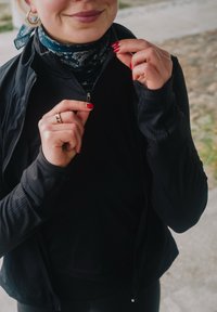Vrouw met roodgelakte nagels die een zwart jasje dichtritst, draagt gouden oorringen, een gouden ring en een blauw patroon sjaaltje om haar nek.