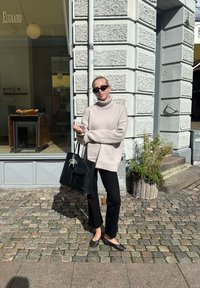 Kremfärgad polokragsweater, svarta byxor, svart toteväska med charm, flatstövlar med prickar, står på en kullerstensgata nära en butik.