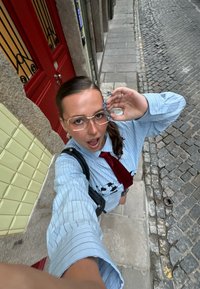 Kék csíkos, bő fazonú ing tehénmintával, piros nyakkendő, aranyszínű szemüveg, hosszú körmök. A háttérben piros ajtó és macskaköves burkolat látható.