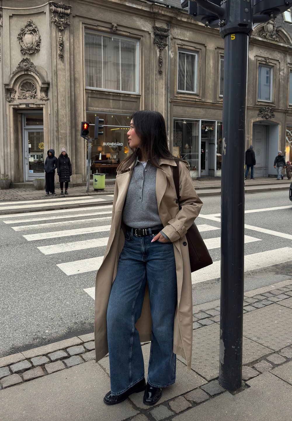 Beige trench coat over a grey polo, paired with wide-leg blue jeans and black shoes. A brown bag is slung over one shoulder.