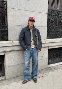 Veste rembourrée sombre, chemise olive, jean bleu clair ample et chaussures marron. Accessoire : sac à bandoulière marron, casquette avec motif du drapeau britannique.