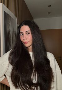 De longs cheveux foncés encadrent un visage maquillé légèrement. La personne porte un pull texturé de couleur claire sur un fond uni.