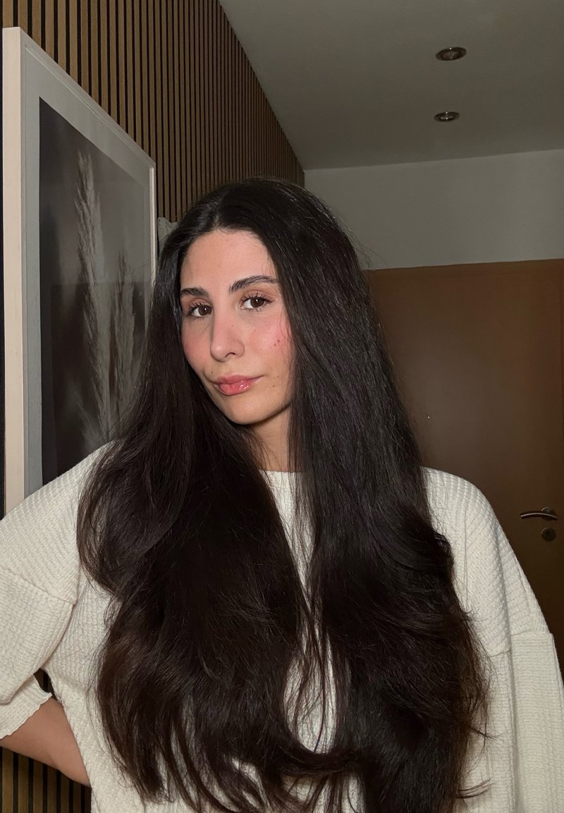 De longs cheveux foncés encadrent un visage légèrement maquillé. La personne porte un pull clair à texture contre un fond uni.