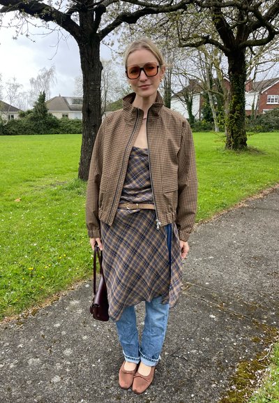 Mujer con chaqueta marrón a cuadros y vestido superpuestos sobre vaqueros azules, de pie en un camino junto a hierba verde y árboles.