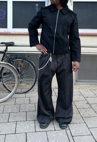 Personne portant une veste noire à fermeture éclair et un pantalon large à fines rayures noir avec un accessoire chaîne, debout à côté de vélos stationnés sur le trottoir.