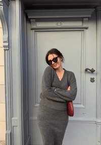 Pull en laine gris tricoté et jupe assortie, avec un col en V profond. Lunettes de soleil noires et sac à main bordeaux complètent le look devant une porte grise.