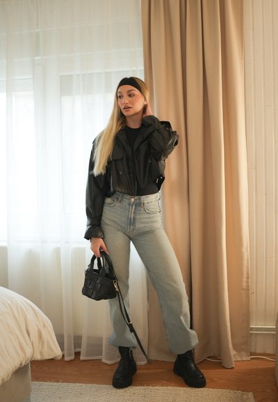 Chaqueta de cuero negra corta sobre una blusa negra, jeans de tiro alto de color azul claro y botines de plataforma negros. Sosteniendo un pequeño bolso tejido negro.