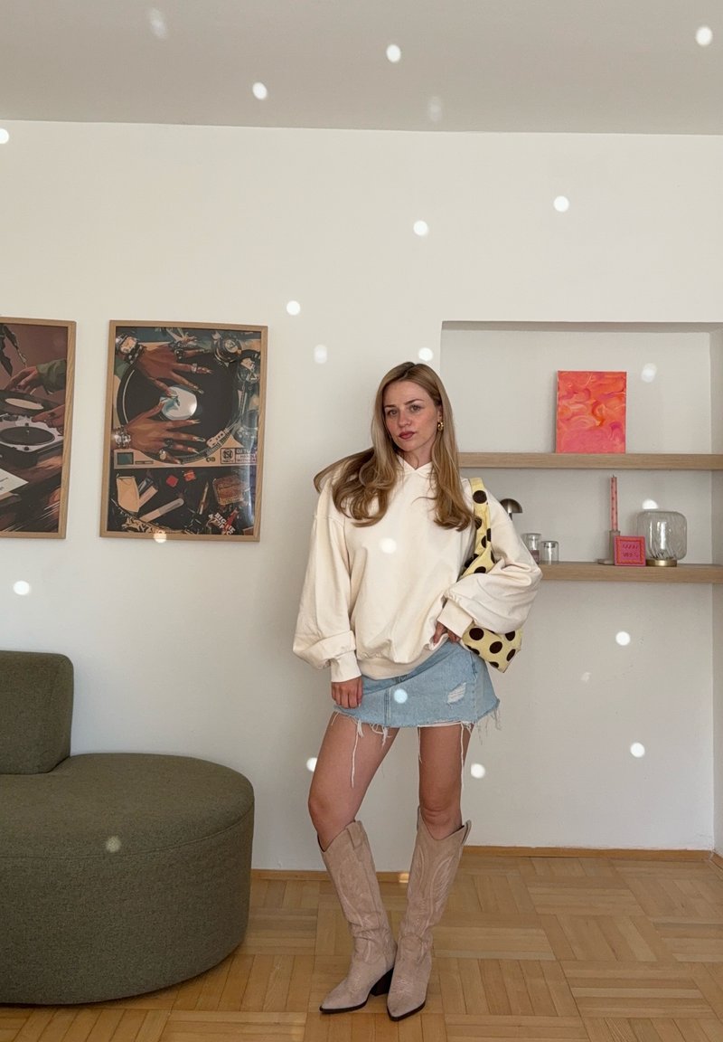 Femme portant un sweat crème oversize, une mini-jupe en denim déchirée, des bottes beiges, tenant un sac à pois dans une pièce moderne avec des œuvres murales et des étagères.