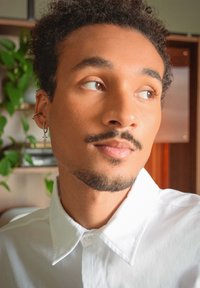 Porträtt av en person med lockigt hår, iklädd en vit skjorta med knappar. Anmärkningsvärda detaljer inkluderar en metallörhänge och en mjuk bakgrund med grönska.