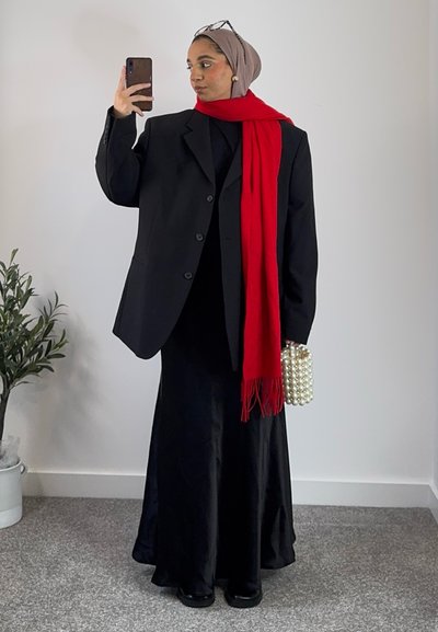 Blazer negro de gran tamaño sobre un vestido negro ajustado, combinado con una bufanda roja brillante y un bolso similar a una perla. Entorno interior con vegetación visible.