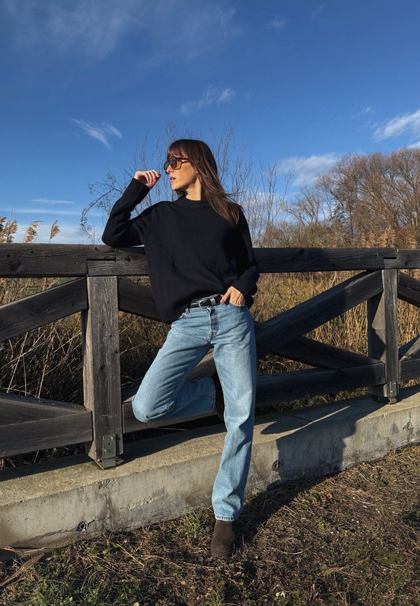 Vrouw met een zwarte trui, blauwe spijkerbroek en zonnebril leunt tegen een houten hek buiten met een heldere lucht en droge vegetatie op de achtergrond.