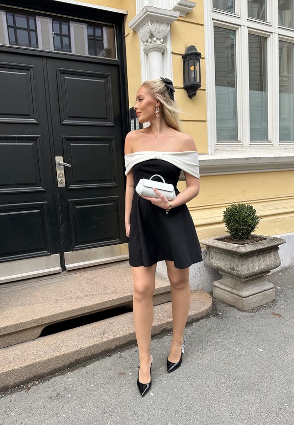 Mustat off-the-shoulder mekko, jossa on valkoinen yksityiskohta, teksturoidusta kankaasta. Mustat kärkitossut, joissa on hopeisia yksityiskohtia. Pitää pientä valkoista käsilaukkua.