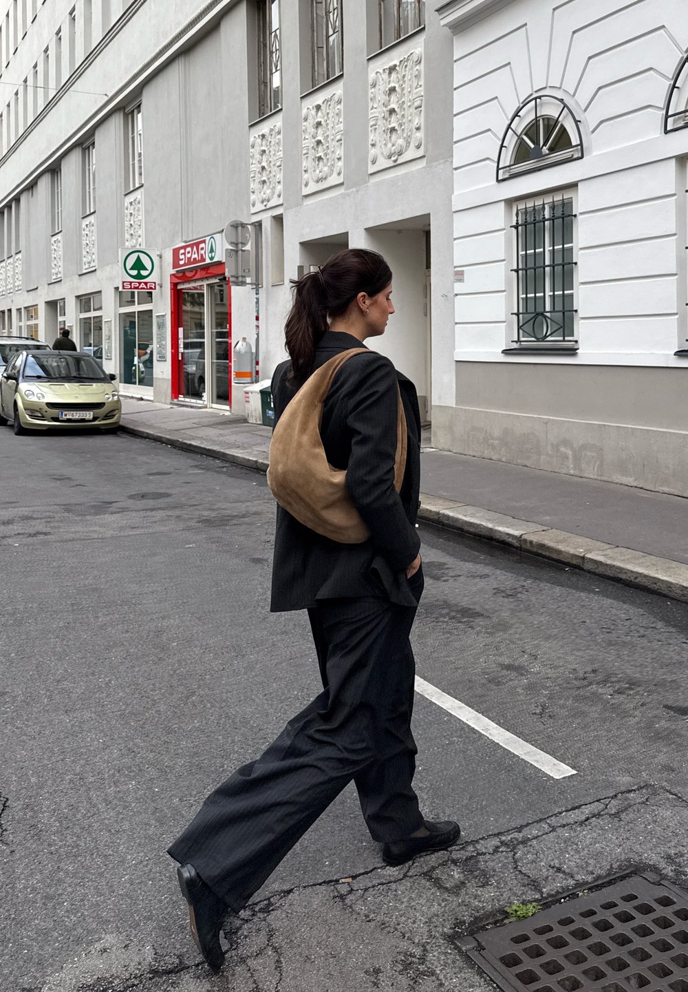 Črne prugaste hlače, črn blazer in torba iz zamsha v svetlo rjavi barvi. Oseba hodi po ulici, v ozadju pa je trgovina Spar.