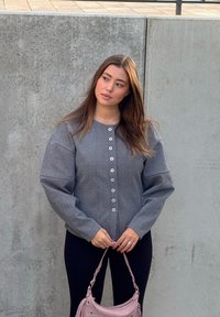 Veste grise boutonnée avec manches oversize, tissu texturé et six boutons. Le mannequin tient un sac à main rose pâle et porte des leggings noirs.