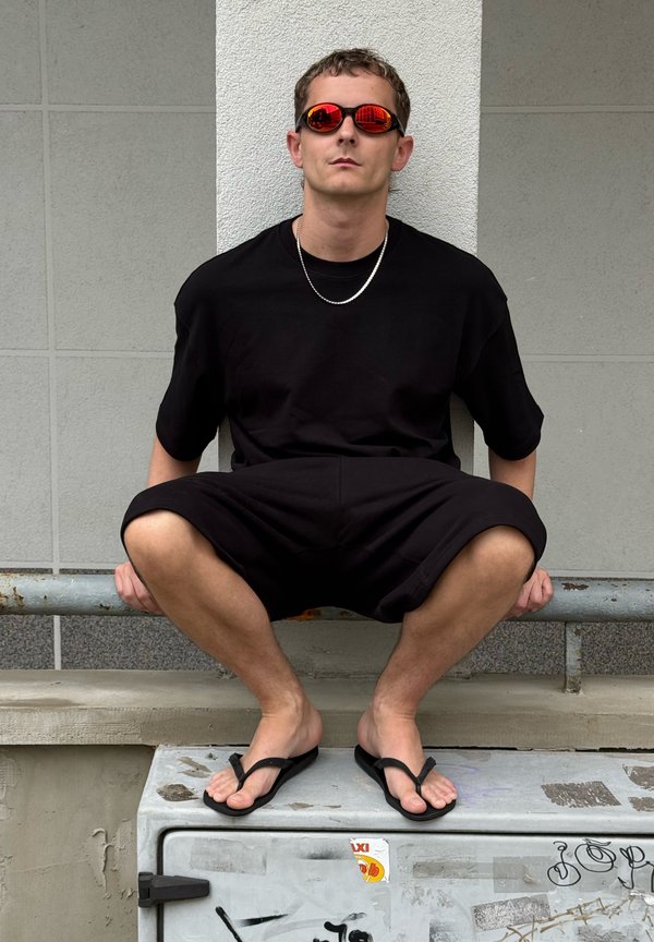 Homme en tenue noire et lunettes de soleil rouges accroupi sur un boîtier de services couvert de graffiti contre un mur carrelé, portant des tongs et une chaîne en argent.