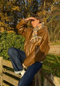 Brun læder bomberjakke med ribbede manchetter og krave, båret over et ternet halstørklæde. Mørk denimjeans og hvide sokker. Model siddende på et træhegn.