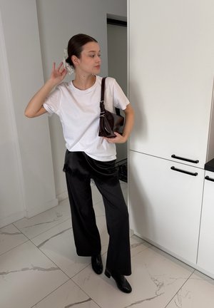 Mujer joven con camiseta blanca, pantalones negros de pierna ancha, botas negras y un bolso oscuro al hombro, arreglándose el cabello con una goma en una habitación blanca.