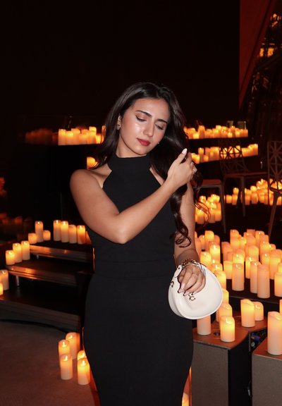 Vestido negro sin mangas con escote alto y silueta ajustada. Mujer sosteniendo un bolso blanco circular. Ambiente de luz suave de vela en el fondo.