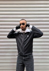 Άνδρας με μαύρο μπουφάν Adidas, τζιν, γάντια και γυαλιά ηλίου ρυθμίζει τα ακουστικά του μπροστά σε κλειστή μεταλλική καταπακτή.