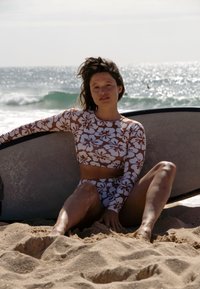 Dugi rukav, kratki top i bikini set u smešnom i belom cvjetnom uzorku, od glatkog, rastezljivog materijala. Postavljen uz surf dasku na pesku.