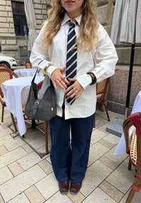 Hvit skjorte med knapper, marineblå stripete slips, brede blå jeans, mørkebrune klomper, grå hobo-veske, gullarmbånd, detaljerte negler med fargeakenter.