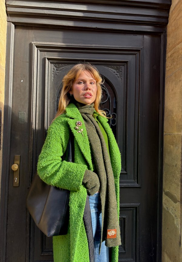 Manteau vert vif texturé avec sac à main noir, écharpe verte et gants. Détail distinctif d'une broche sur le manteau. Jean en denim visible. Fond de porte noire.