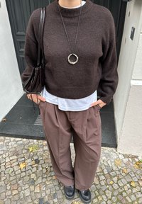 Person, die einen dunkelbraunen Pullover, ein weißes Unterhemd, lockere braune Hosen, schwarze Schuhe, eine silberne Ketten mit Kreisanhänger und eine dunkelbraune Umhängetasche trägt, steht auf Kopfsteinpflaster.