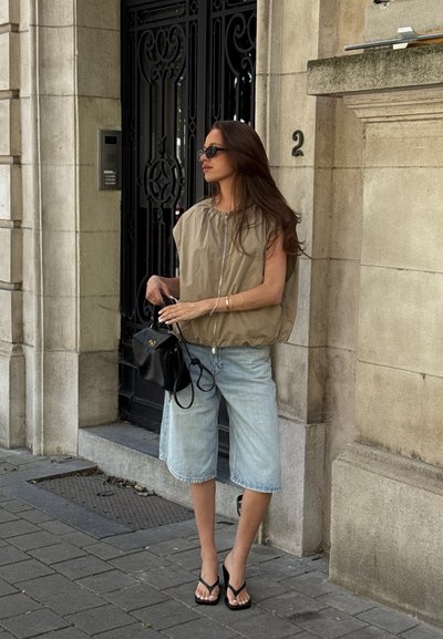 Chaleco acolchado beige con cremallera frontal, pantalones cortos de mezclilla claros, bolso negro y chanclas, de pie contra una pared de piedra y una puerta ornamentada.