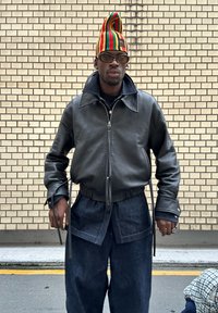 Svart läderjacka med dragkedja och bred krage, insvängd midja; lösa mörk denimbyxor. Mångfärgad randig hatt, transparenta glasögon, silverringar.