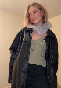 Veste noire en cuir surdimensionnée, top à bretelles fines vert olive avec ornements floraux, écharpe grise en tricot, pantalon noir ajusté.