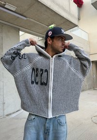 Juodai baltas striukės užtrauktukas su plačiomis pečiais, pasižymintis oversized siluetu ir ryškiais tekstiniais akcentais. Kombinuojama su šviesiai mėlynomis džinsinėmis kelnėmis.