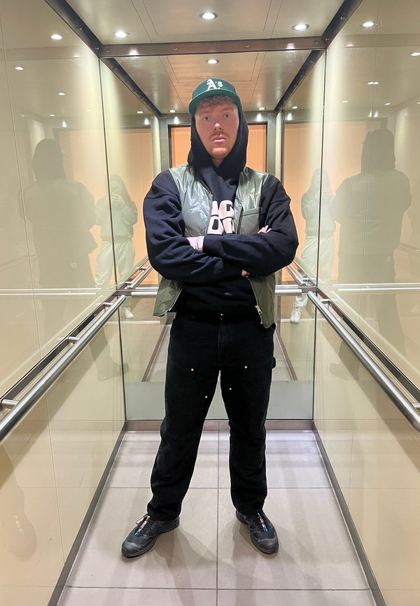 Green padded vest over a black hoodie, black pants, and black shoes. Captured in a mirrored elevator with bright lights.