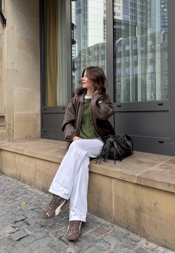 Braune Bomberjacke über einem grünen Knopf-Front-Pullover, weiße weite Hosen und braune Sneakers. Texturiertes schwarzes Handtasche auf dem Boden.