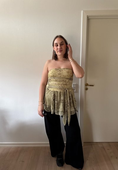 Mujer de pie en el interior, llevando un top amarillo sin tirantes con estampado y fajín, y pantalones negros de pierna ancha, arreglándose el cabello con una mano.