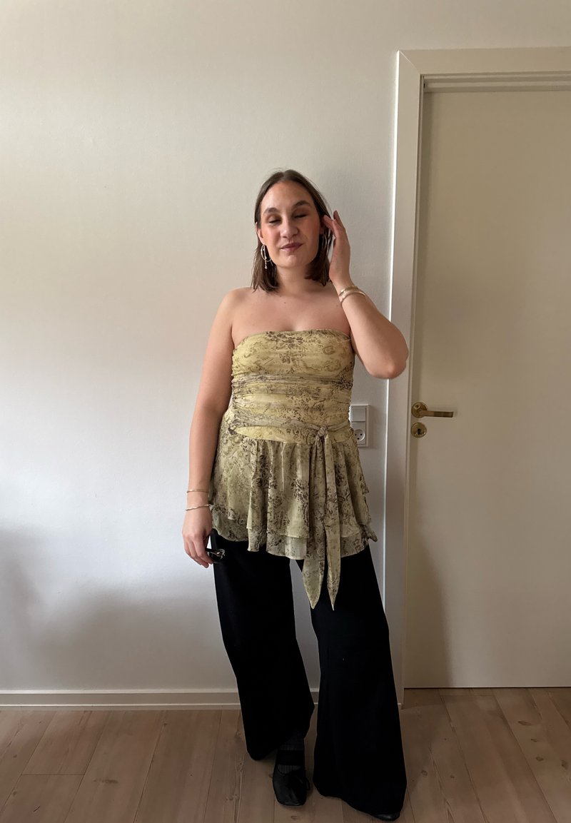 Woman standing indoors wearing strapless patterned yellow top with sash and black wide-leg pants, adjusting hair with one hand.