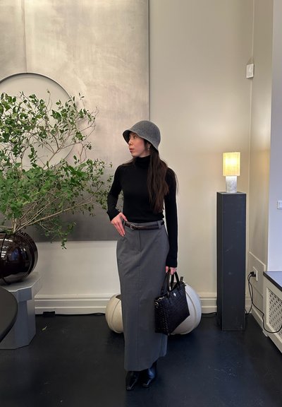 Mujer con falda gris, blusa de manga larga negra y sombrero gris, de pie en el interior sosteniendo un bolso negro junto a una planta grande y una lámpara encendida.