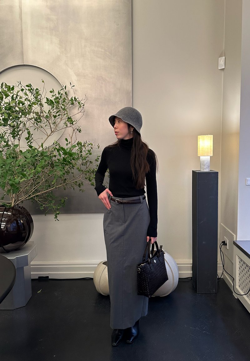 Vrouw in een grijze rok, zwart shirt met lange mouwen en een grijze hoed staat binnen, houdend een zwarte handtas naast een grote plant en een brandende lamp.