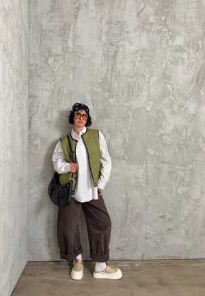 Persona con chaleco acolchado verde oliva sobre camisa blanca, pantalones marrones de pierna ancha, zapatos crema, bandana negra y gafas de sol naranjas, de pie en interiores.