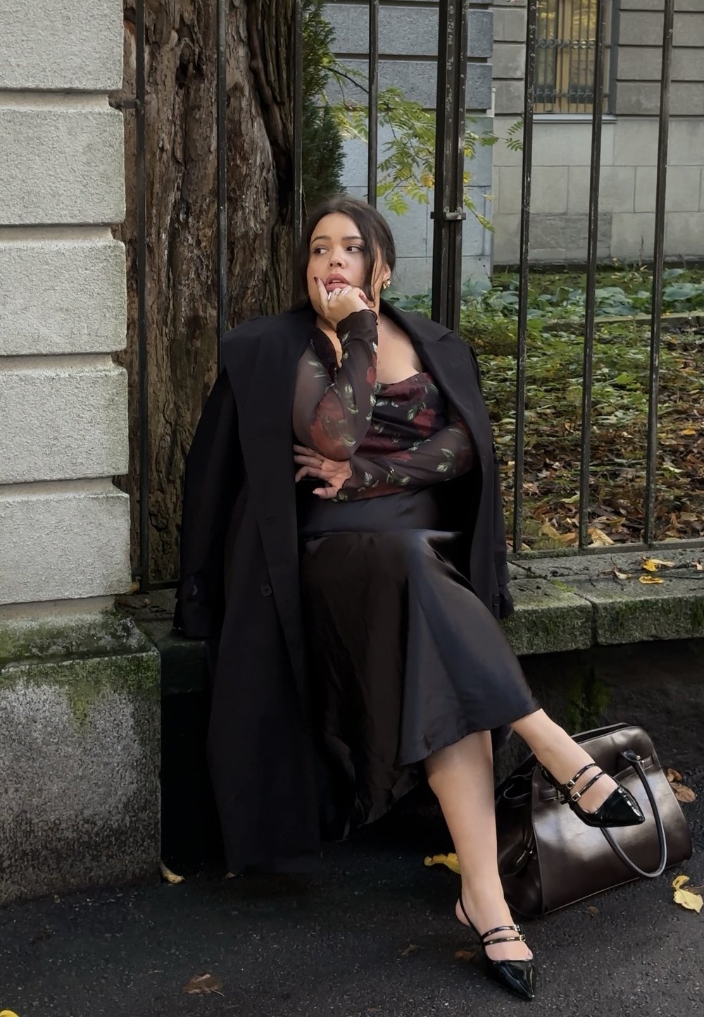 Zwarte satijnen slipdress gecombineerd met een doorschijnend bloemen top en een lange jas, aangevuld met black hakken met bandjes en een bruine handtas.